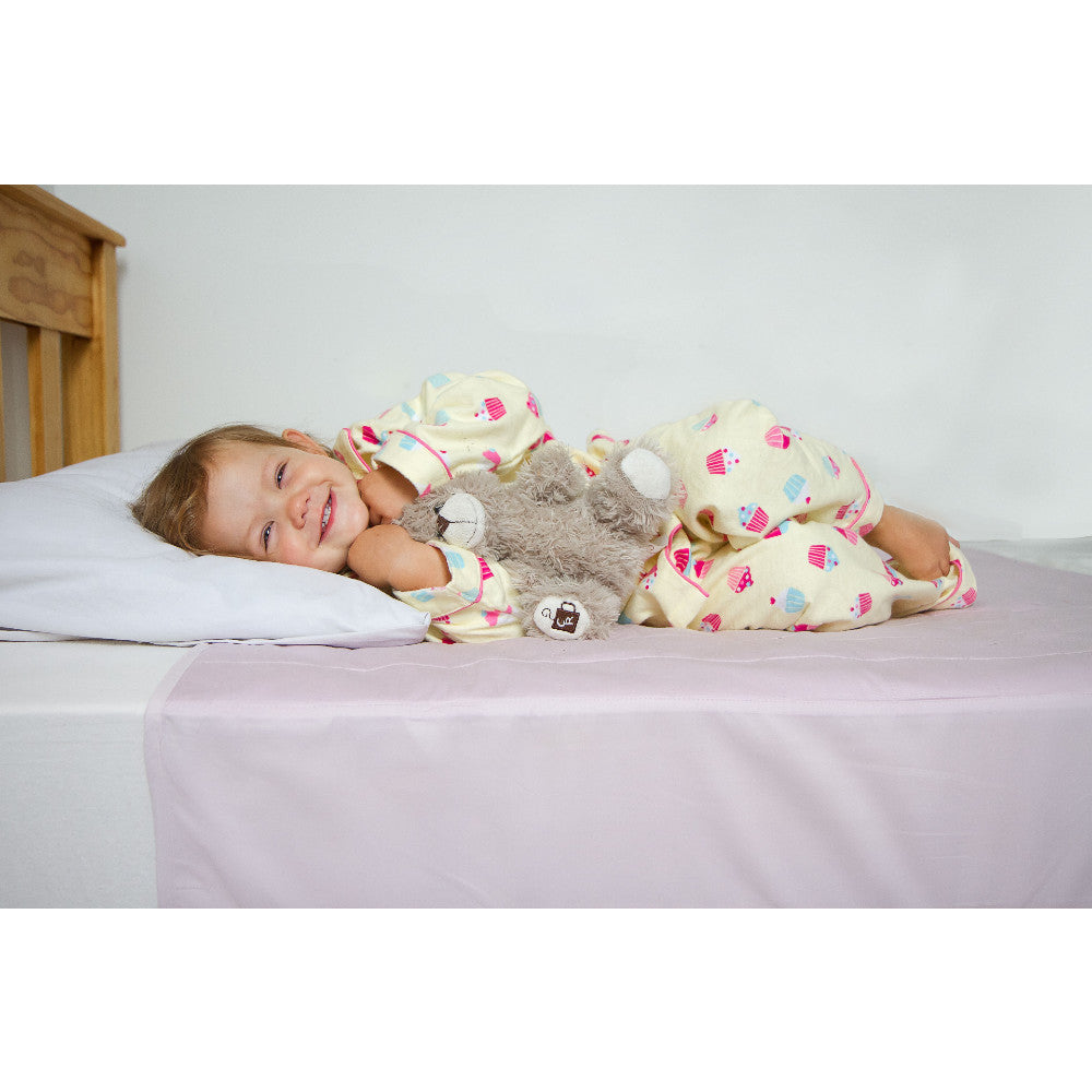 A smiling toddler lies on a bed wearing yellow pyjamas with a colourful cupcake print, holding a gray stuffed animal. The room has a white wall and a light wood headboard, with a pale pink brolly sheet protector covering the bed.