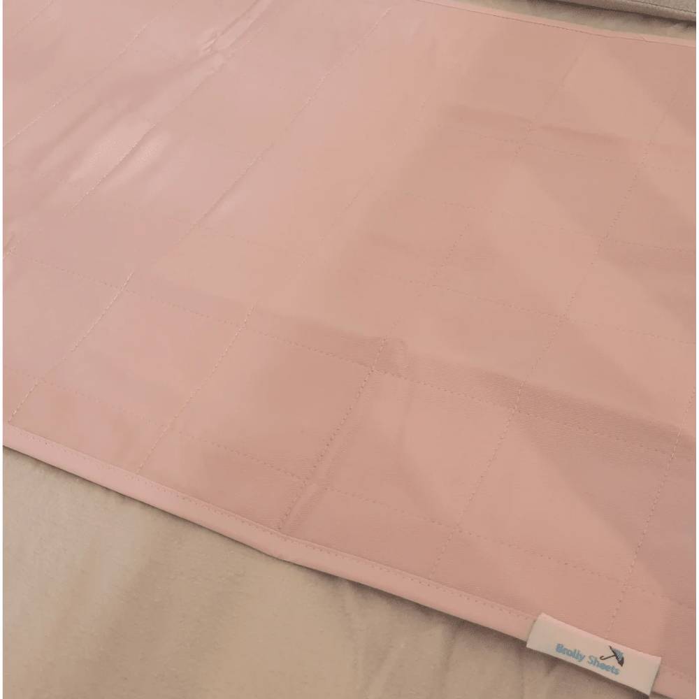 A close-up of a light pink quilted brolly sheet with a stitched grid pattern, laid flat on a neutral beige surface. A small white fabric tag with blue text is visible in the bottom right corner.