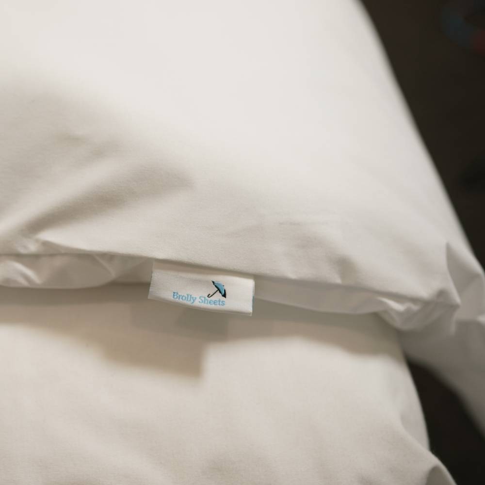 A close-up of a white pillowcase with a small fabric tag reading 'Brolly Sheets' and a small blue logo, resting on a white sheet.