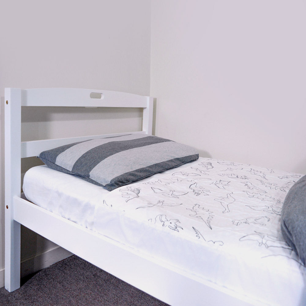 A white wooden single bed frame with a white quilted brolly sheet featuring black dinosaur line drawings, a gray and white striped pillow, and a gray blanket, set against a plain light wall.