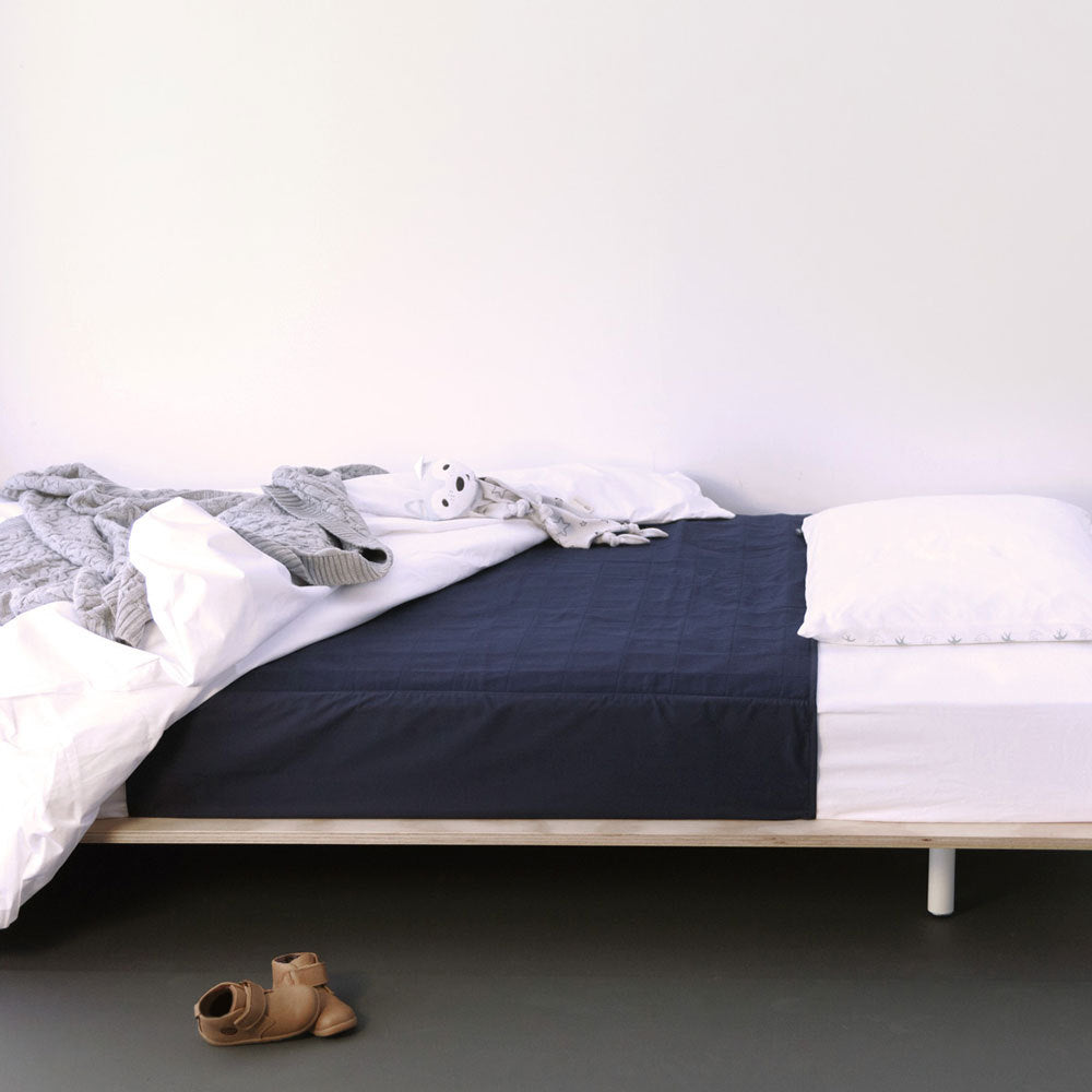 A single bed with a light wood frame, dressed in a dark navy quilted brolly sheet pad, white sheets, and a gray knit blanket, with a small stuffed animal and a pair of tan boots on the floor.