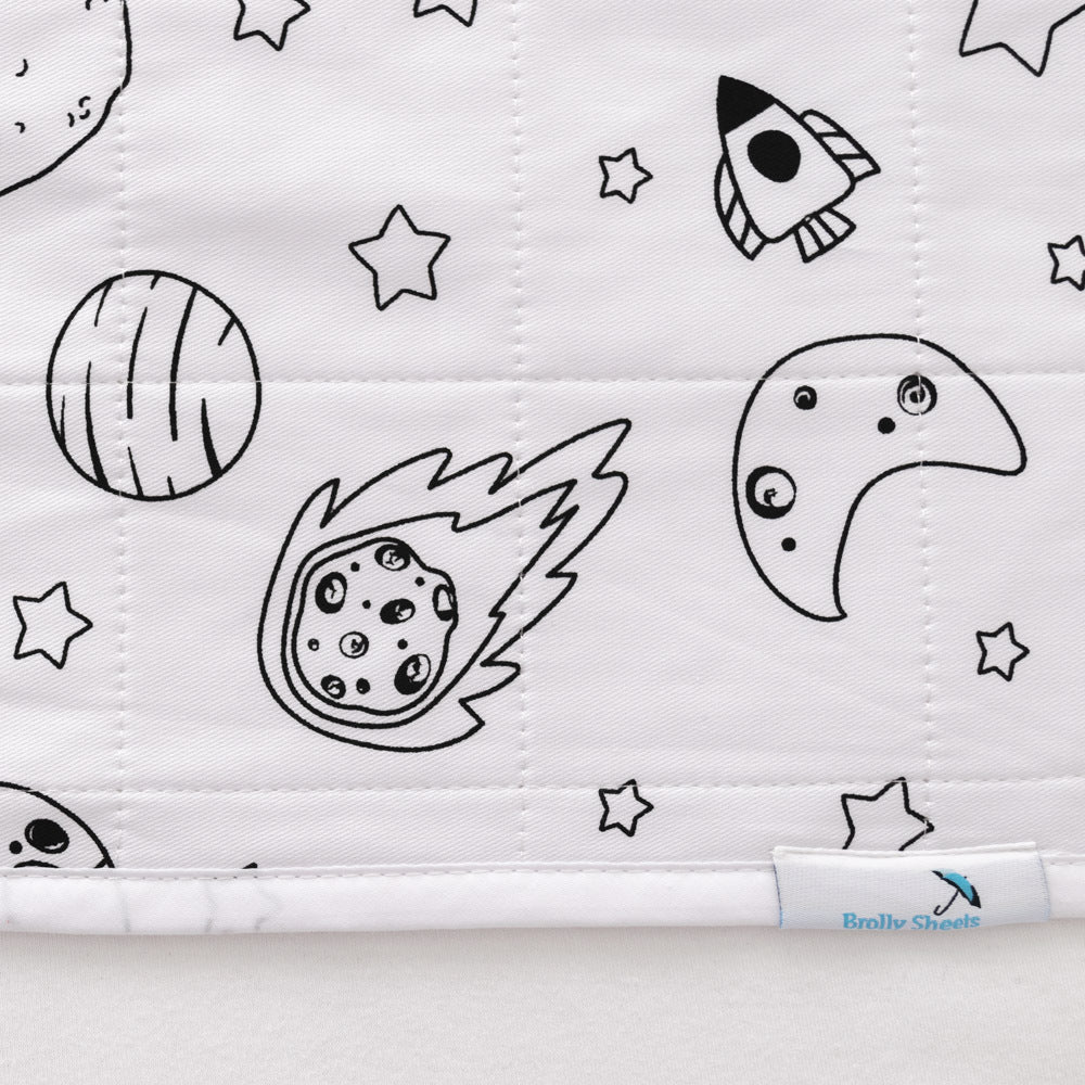 Close-up of a white quilted waterproof ped pad featuring a black line art space print with stars, planets, a moon, and a rocket. A small fabric tag at the bottom right reads 'Brolly Sheets'