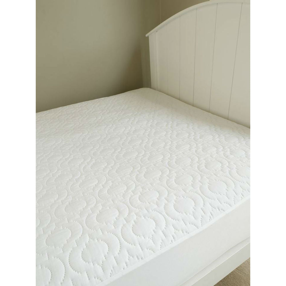 A brolly sheet white waterproof quilted mattress topper with pattern, shown on a bed with a white wooden headboard against a neutral beige wall.