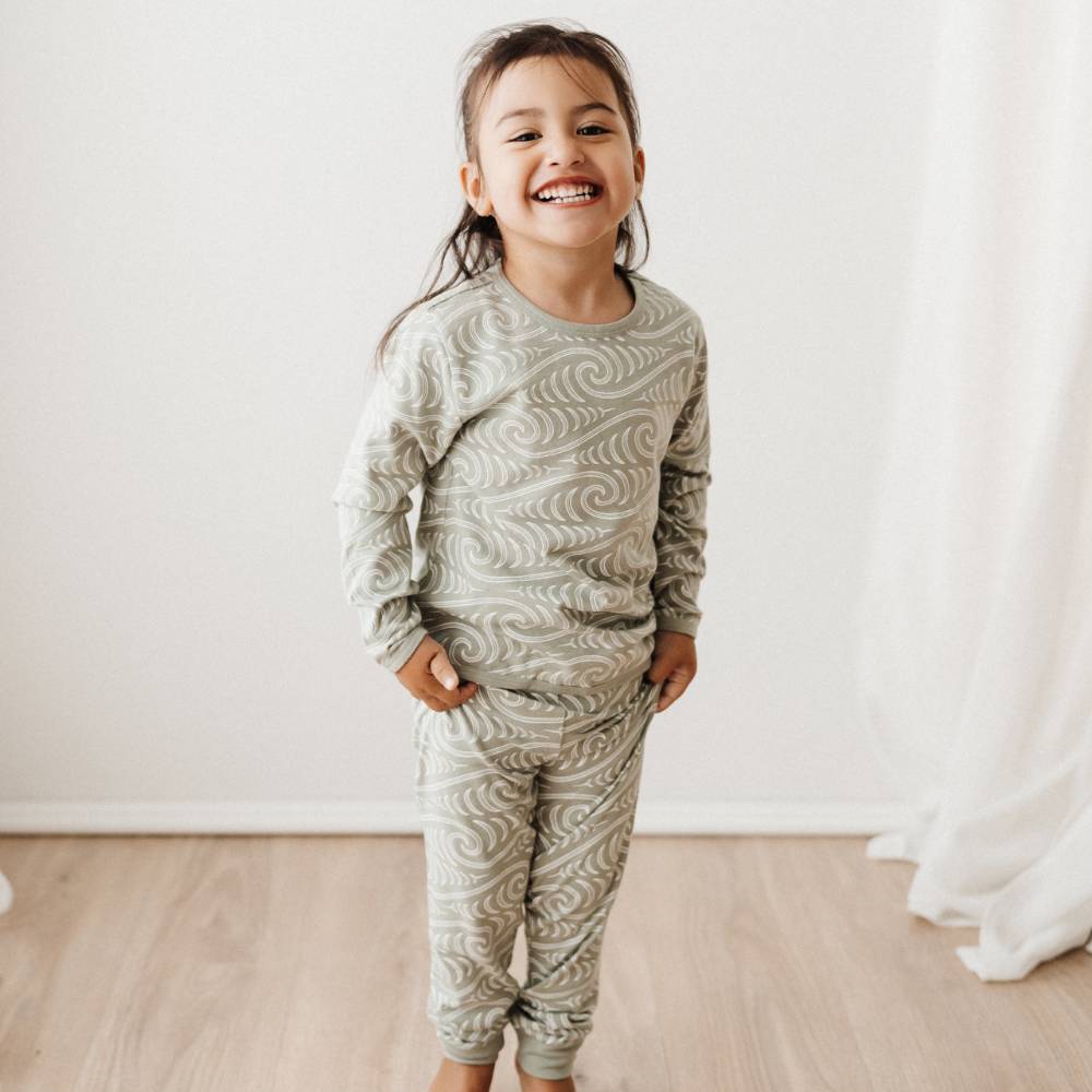 Child wearing a maori patterned pajama set standing in a room with a white wall and wooden floor.