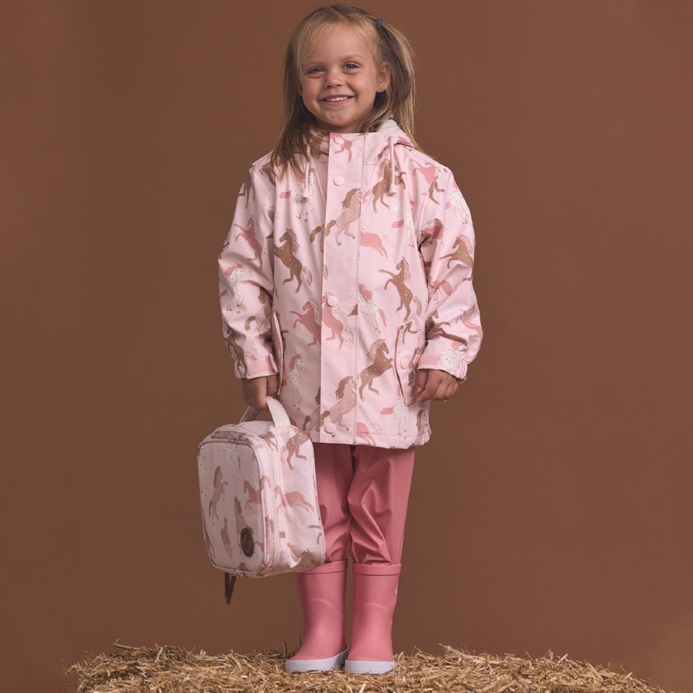 Child wearing a pink raincoat with horse pattern, holding a matching lunch bag, on a brown background