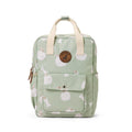 A light sage green backpack with a white apple pattern, featuring cream-colored top handles, a front pocket with a snap button, and a circular brown leather patch on the front.
