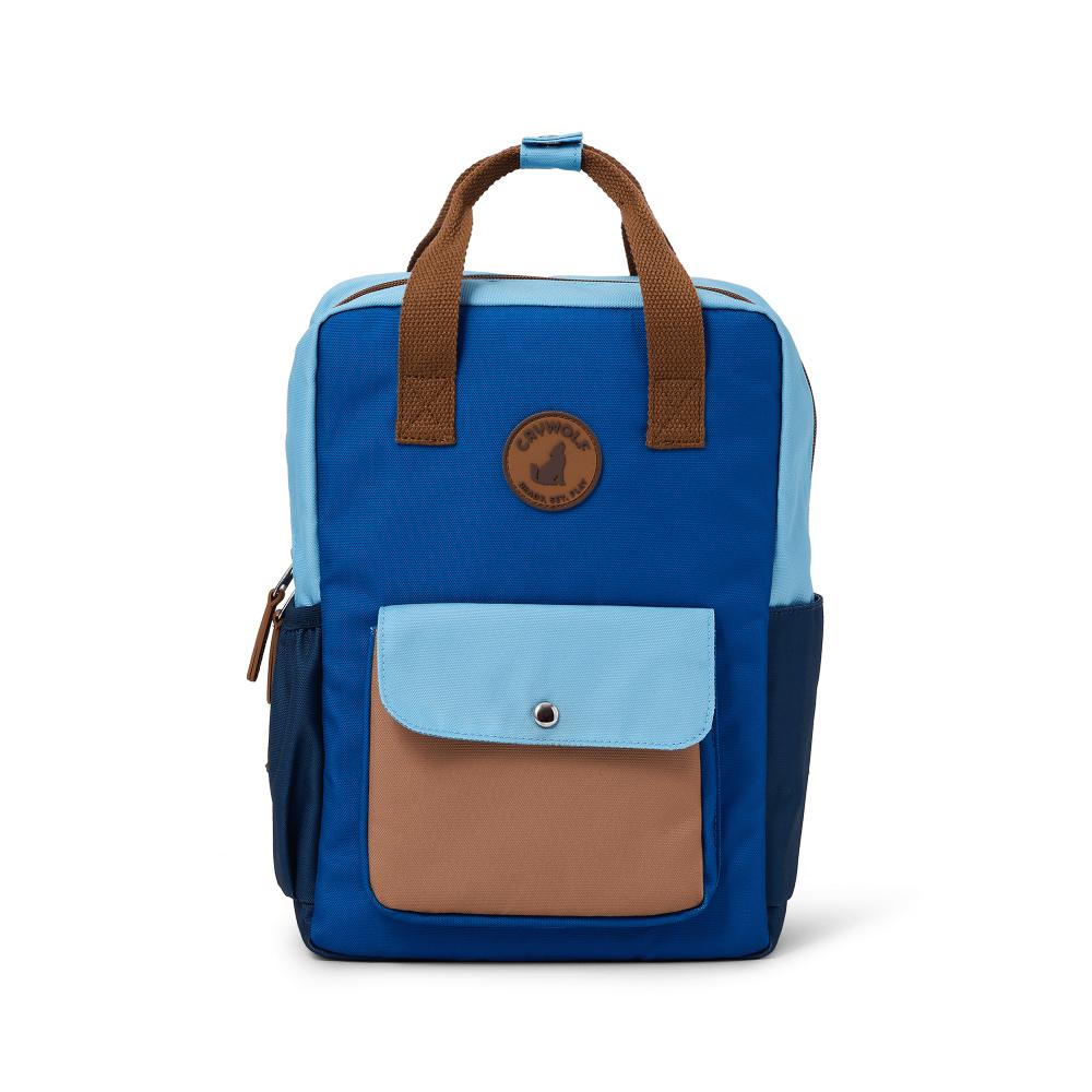 A color-blocked backpack in blue, light blue, and brown, featuring dual top handles, a front pocket with a snap closure, and a circular brown logo patch on the upper front.