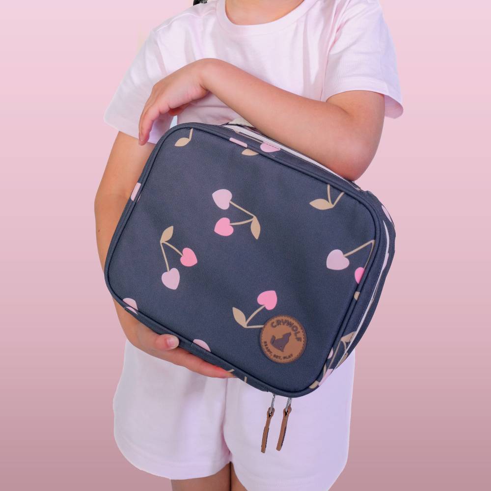 A child wearing a white t-shirt and shorts holds a navy blue zippered lunch bag with a pink and beige cherry pattern. The bag features a circular brown patch with the brand name 'CRYWOLF'.