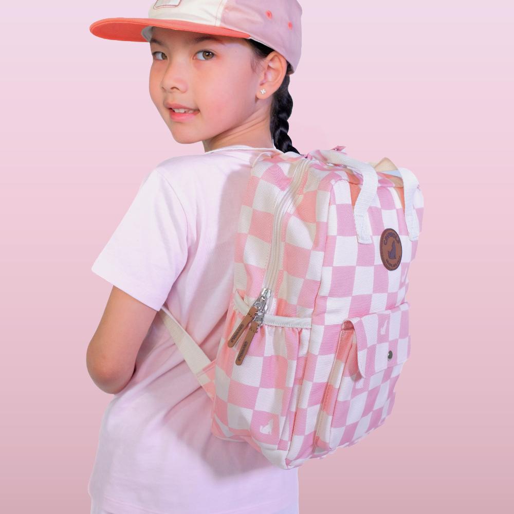 A child wearing a pink and white checkered backpack, a pink and white baseball cap, and a light pink t-shirt, shown from a three-quarters back view against a solid pink studio background.