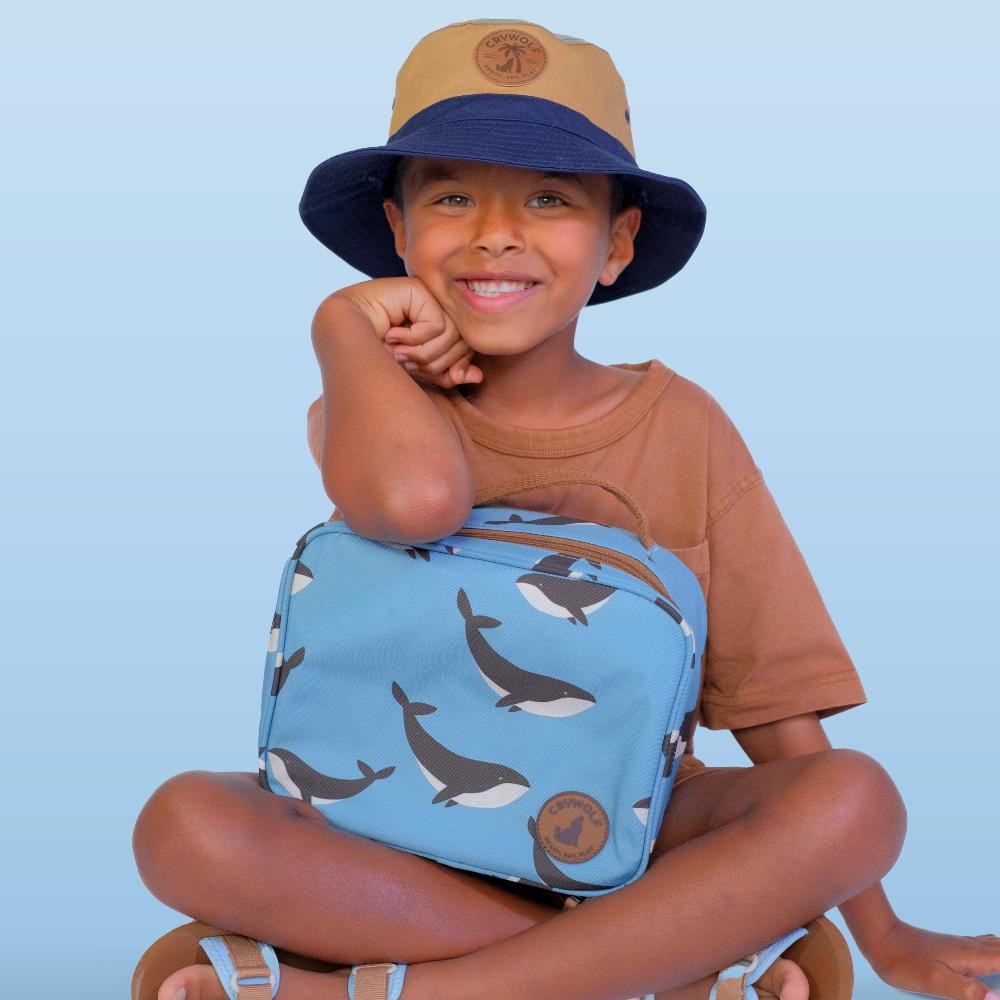 A smiling young boy sits cross-legged against a light blue background, wearing a tan and navy bucket hat, a brown t-shirt, and holding a light blue whale-print lunch bag.
