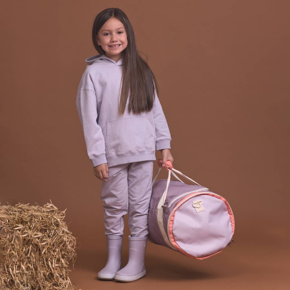 A young girl with long dark hair stands against a solid brown studio background, wearing a matching light lavender sweatshirt and jogger pants set with matching rain boots. She is holding a lavender cylindrical duffel bag with peach trim and cream handles.