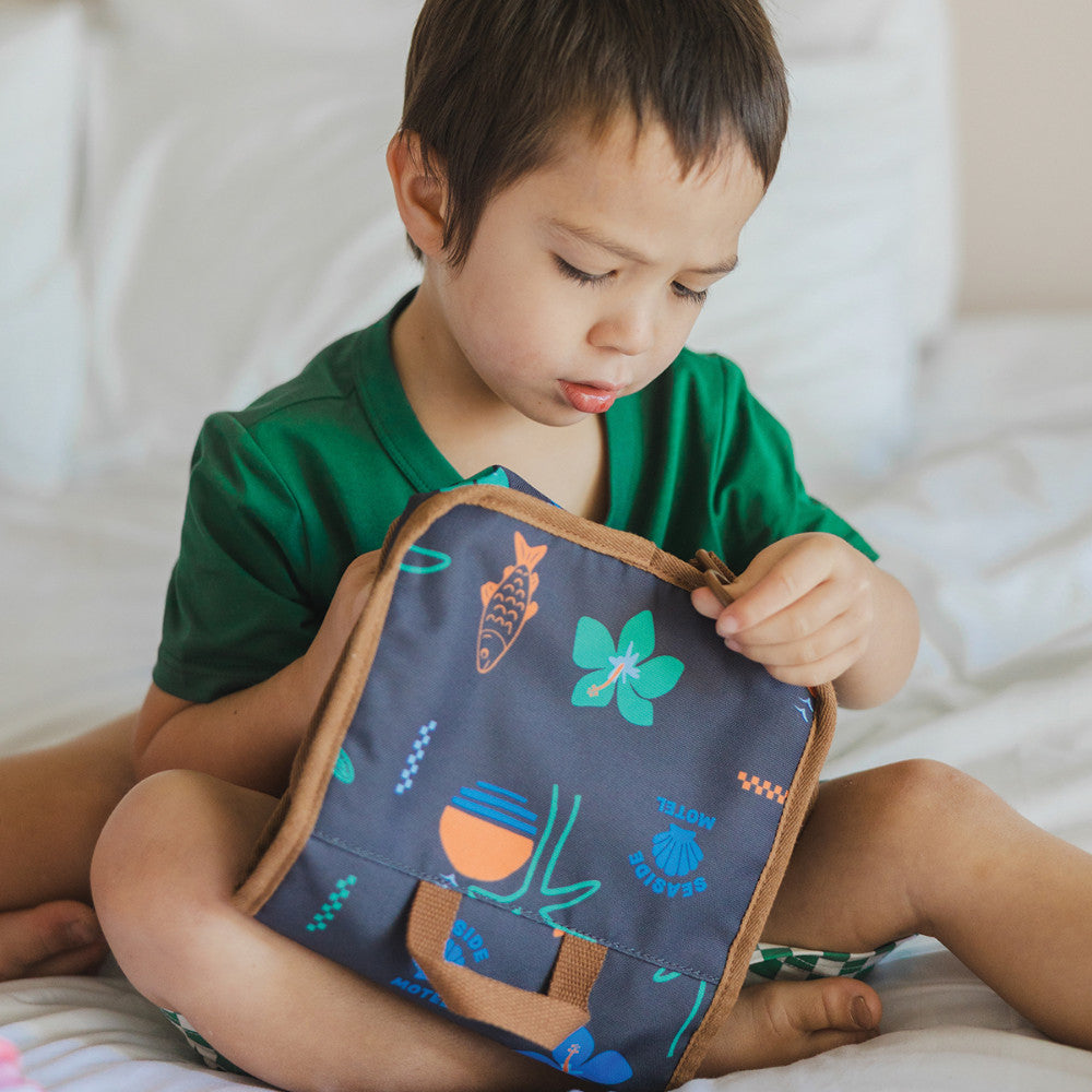 A young child sitting on a white bed holding a navy blue toiletry bag with a tropical fish and flower pattern.