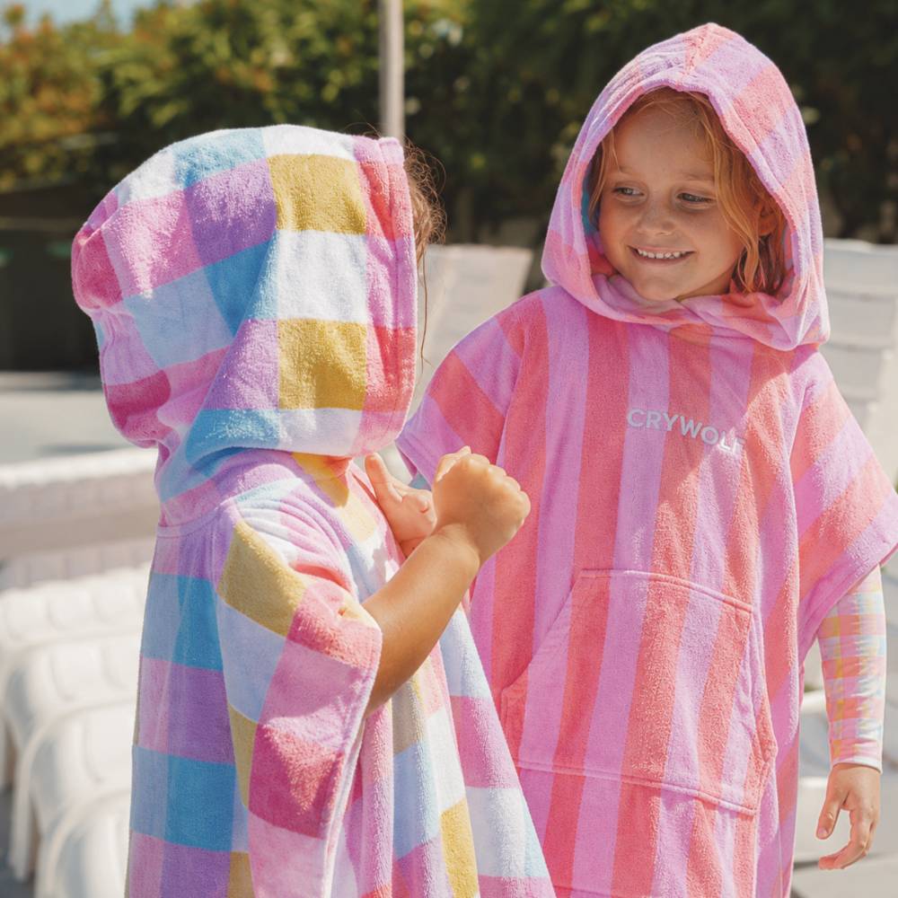 Two children wearing Crywolf hooded beach towels in a sunny outdoor setting. One child is facing away, showing a pastel multicolor checkered pattern in pink, blue, yellow, and white.