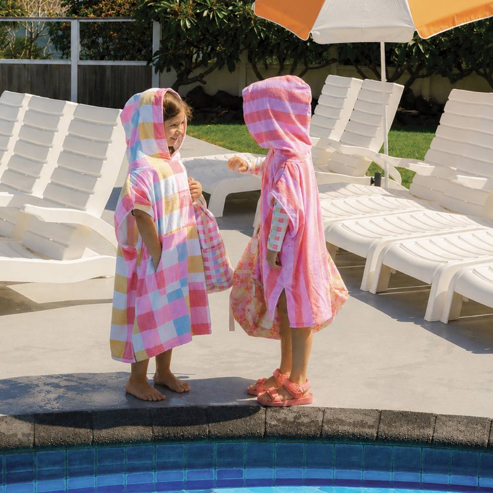 Two children standing by a swimming pool wearing pink and peach checkered hooded towels, with white lounge chairs and an umbrella in the background.