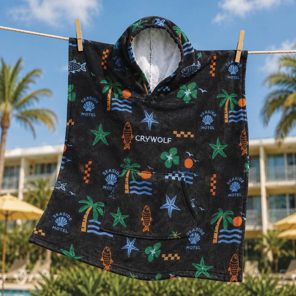 A black hooded towel with a colorful tropical beach print hangs from wooden clips on a rope outdoors, with palm trees and a resort building in the background. The print includes palm trees, starfish, waves, seashells, fish, and the text 'CRYWOLF' and 'SEASIDE MOTEL'.
