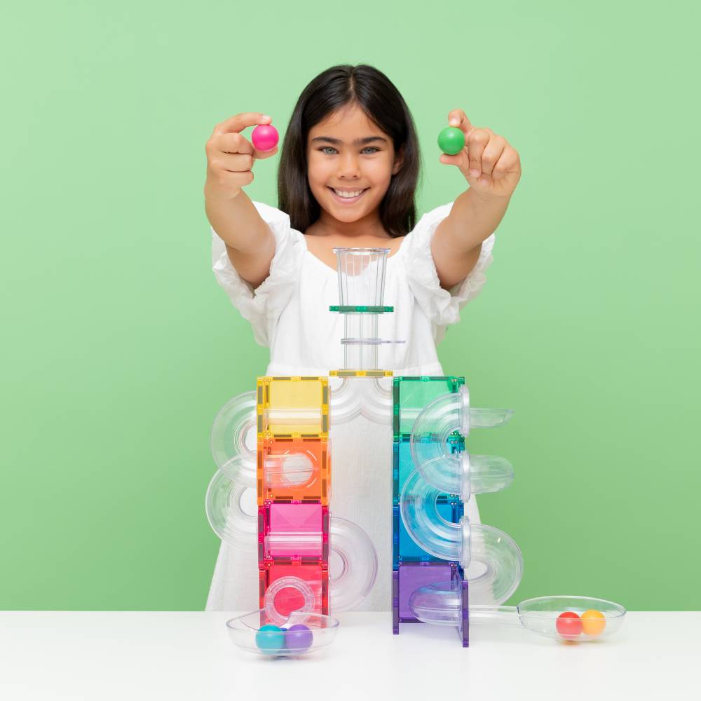 A smiling young girl in a white dress holds up two colorful marble balls, one pink and one green, above a colorful marble run toy set on a white table against a light green background.