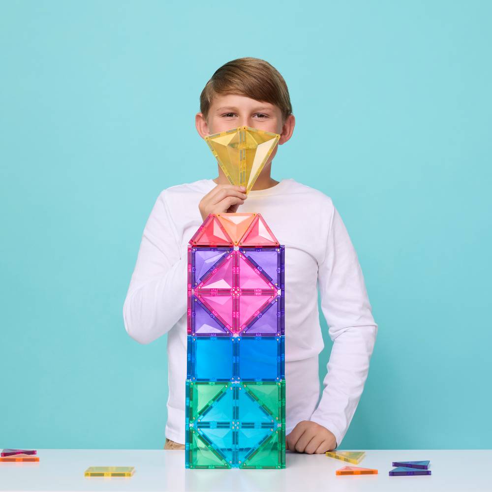A young boy wearing a white long-sleeve shirt stands behind a colorful magnetic tile tower, holding a yellow geometric tile piece in front of his face. The tower is constructed from translucent, rainbow-colored magnetic tiles on a white table against a solid light blue background.