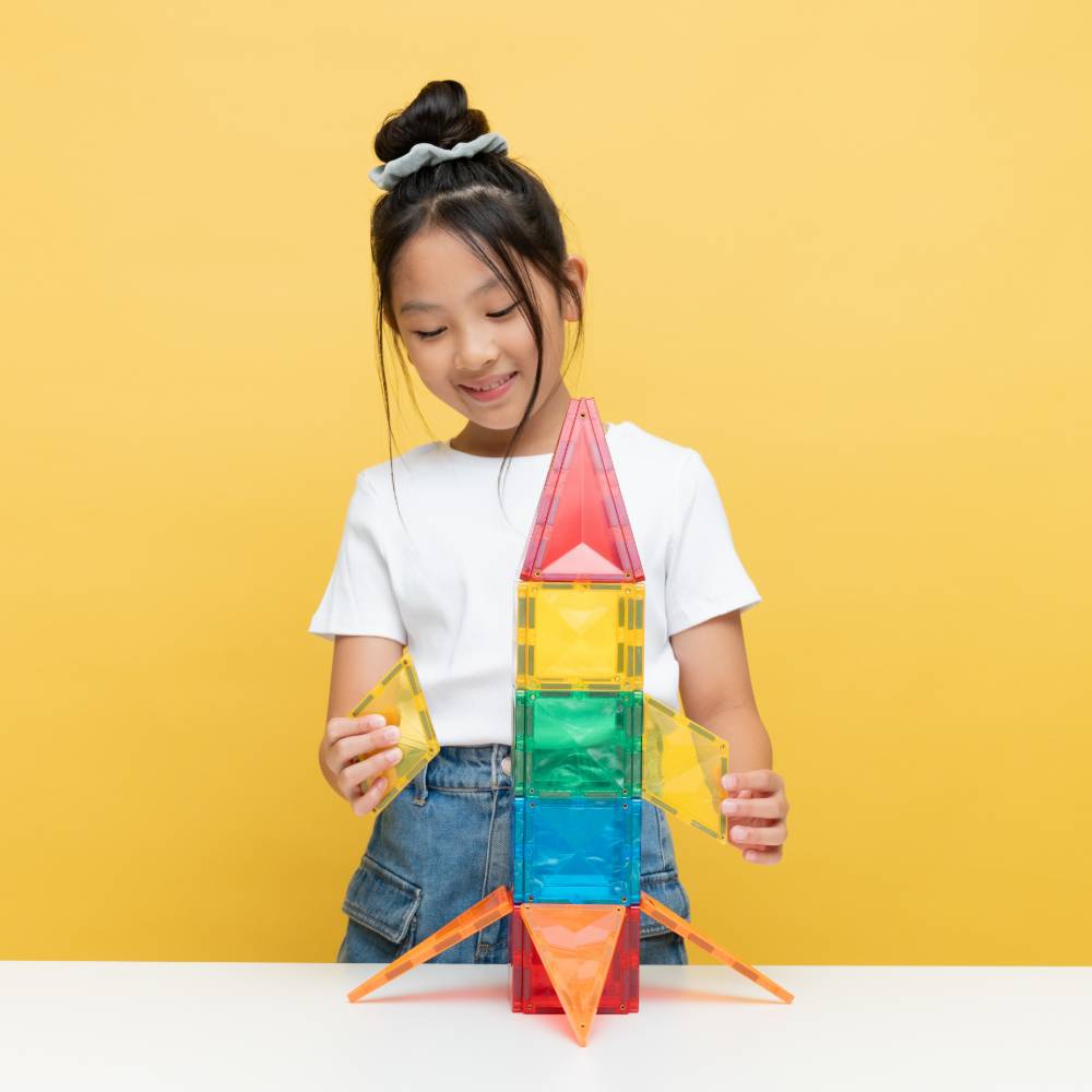 A young girl with her hair in a bun is smiling and building a colorful translucent plastic rocket ship with magnetic building tiles on a white table against a solid yellow background.