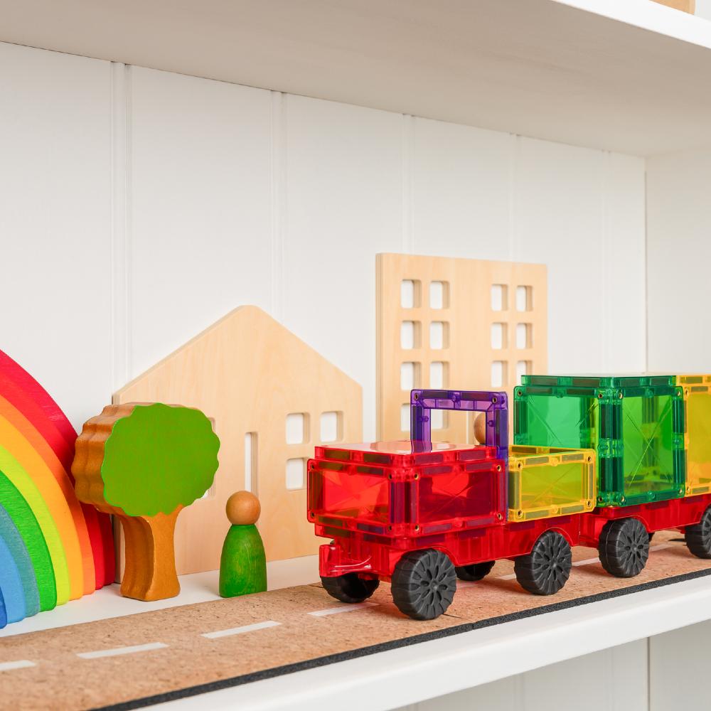 A colorful children's magnetic tile train set on a shelf, with translucent red, green, yellow, and purple train cars and black wheels, displayed beside wooden house shapes, a rainbow, and toy figures.