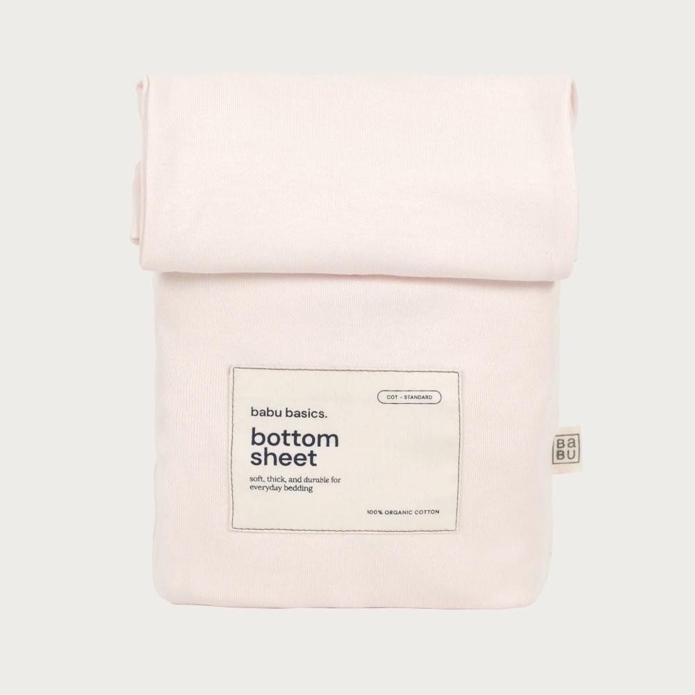 A pale blush pink cotton bottom sheet in a roll-up canvas style, featuring a central white label that reads 'babu basics. bottom sheet' and a small side tag, shown on a light gray background.