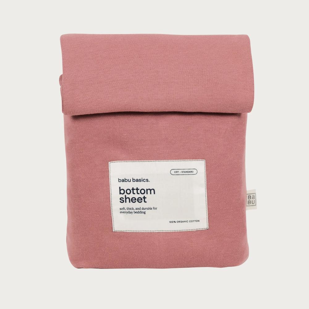 A dusty rose pink cotton bottom sheet by babu basics, neatly folded and rolled up with a white product label on the front, set against a solid off-white background.
