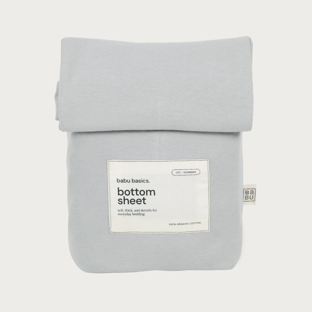 A folded light gray cotton bottom sheet with a white paper label reading 'bubu basics bottom sheet' and a small side tag, photographed on a clean off-white background.