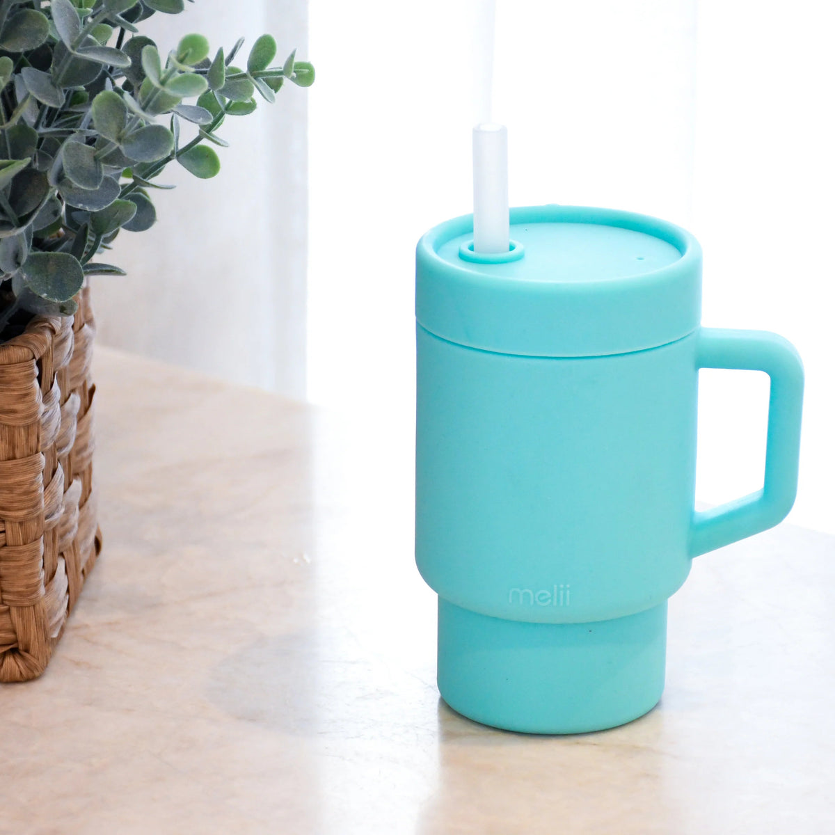 Turquoise children's trainer mug with a straw on a light surface next to a plant