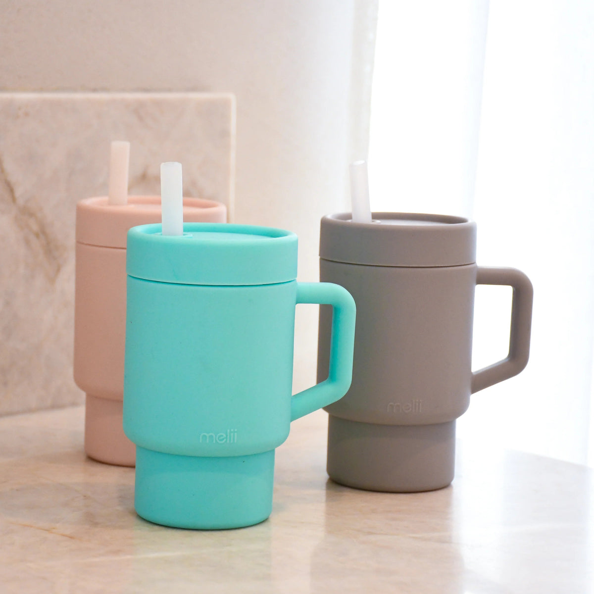 Three Melii silicone kids tumblers with handles on a light surface, one with a straw.