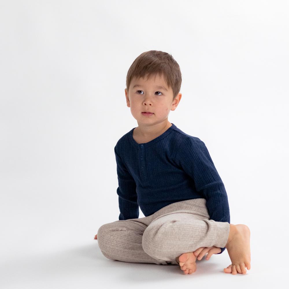 Child sitting cross-legged on a white background wearing a navy sweater and beige woolbabe pants.