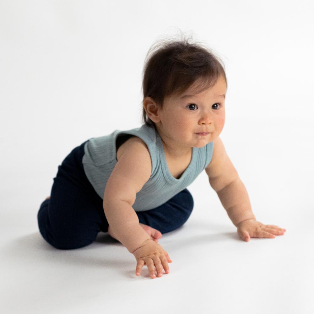Baby crawling on a white surface wearing a light blue sleeveless bodysuit and dark pants.