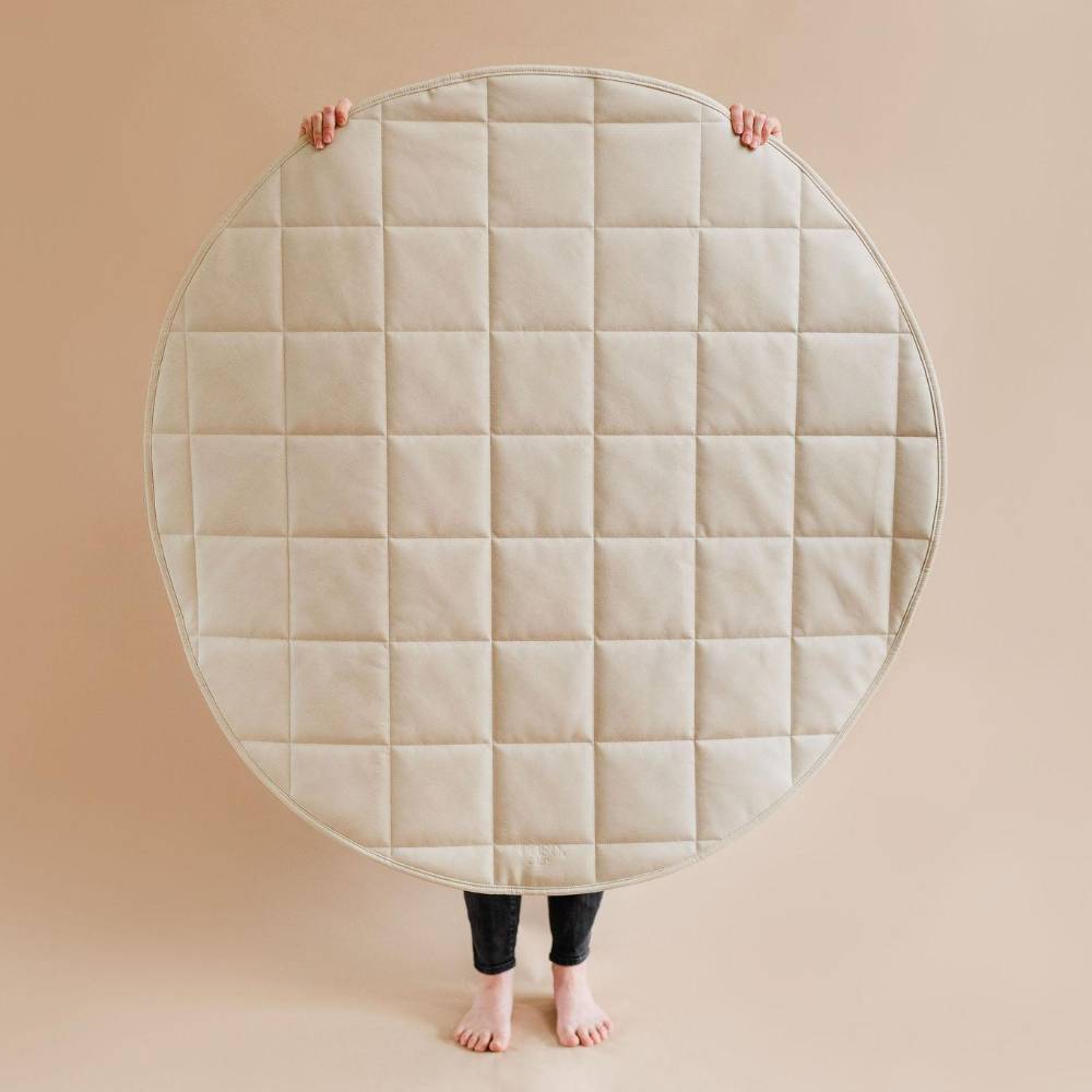 A person holding up a very large round quilted mat or pad in a studio setting. The product is light cream or off-white with a stitched square grid pattern.