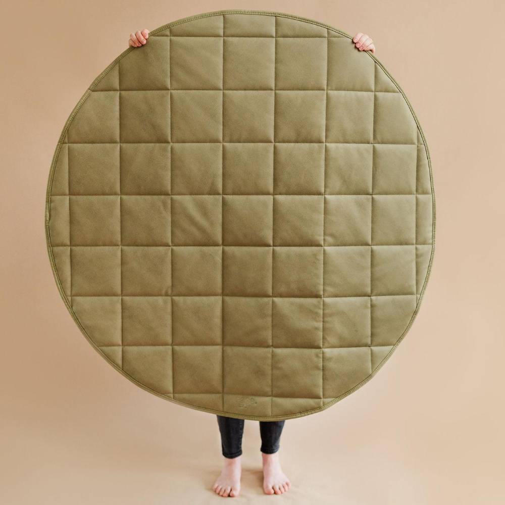 A large round quilted floor mat or play mat in an olive green color, held up by a person whose legs and bare feet are visible against a solid beige background.