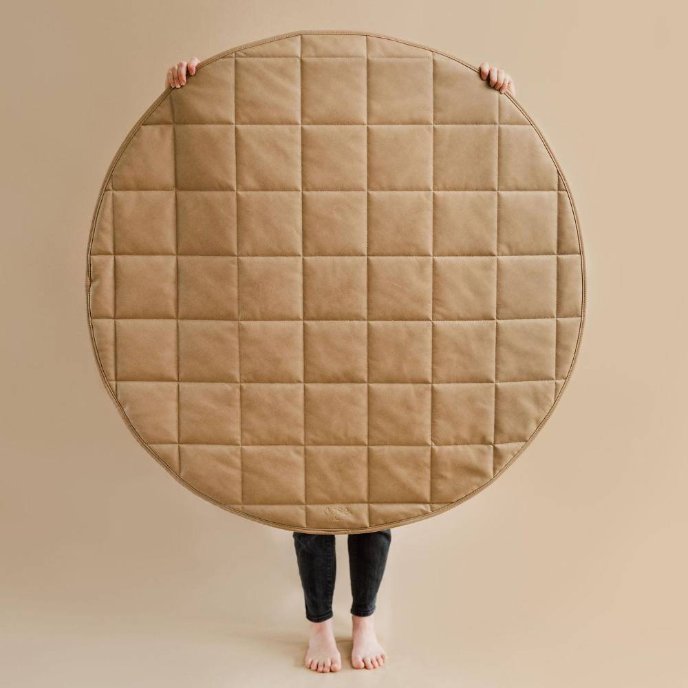A large round quilted tan floor mat or rug held up by a person whose hands and bare feet are visible against a plain beige background.