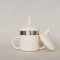 An off-white stainless steel sippy cup with a matching lid resting beside it, featuring a stainless steel band around the middle and a small handle on the left side, set against a neutral light gray background