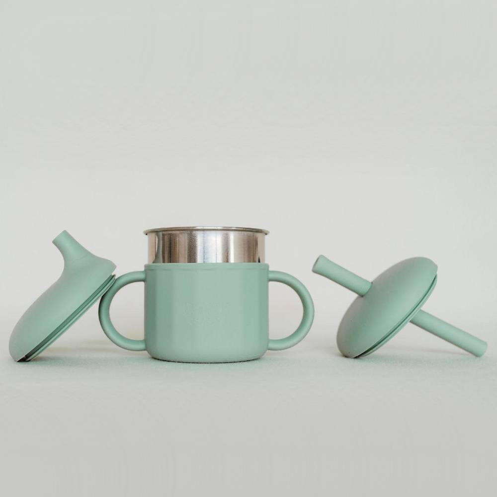 A mint green silicone baby cup and lid set with a stainless steel inner cup, displayed on a light gray background.
