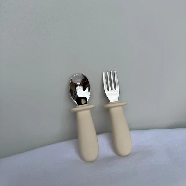 A baby spoon and fork set with matte beige handles and polished silver metal heads, standing upright on a white textured surface against a light gray wall.