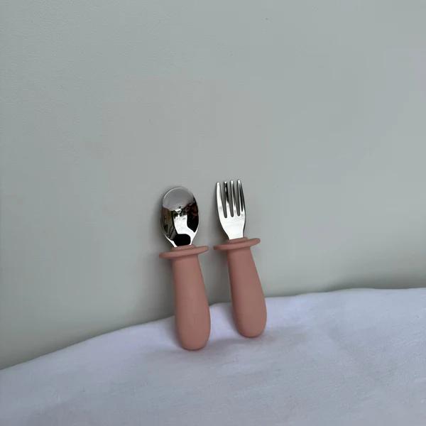 A child's silicone utensil set with a spoon and a four-tine fork, featuring dusty pink handles and silver metal heads, resting on a white surface against a light gray wall.
