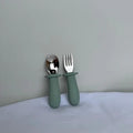 A baby utensil set featuring a spoon and a fork with matte sage green silicone handles and polished stainless steel heads, standing upright against a neutral gray background.