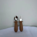 A baby feeding utensil set consisting of a spoon and a fork with matte brown silicone handles and silver metal heads, standing upright on a light gray fabric surface against a matching gray wall.