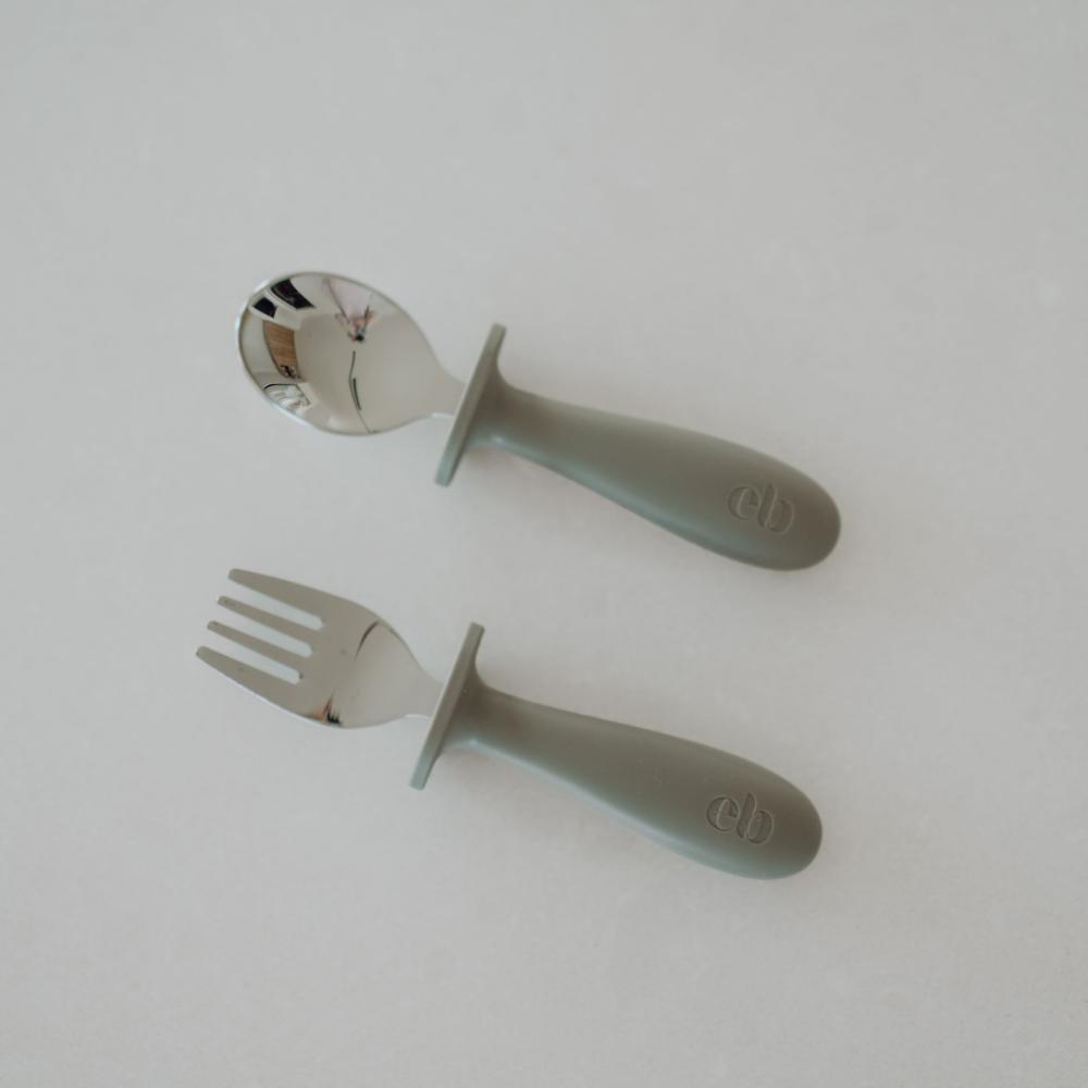 A baby feeding set consisting of a silicone spoon and fork with silver stainless steel tines and handles, displayed on a light gray background.