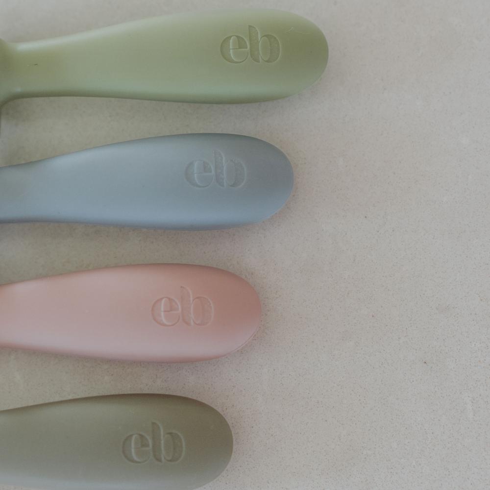 Four pastel-colored silicone baby spoons with embossed 'eb' logos, arranged vertically on a neutral beige background.