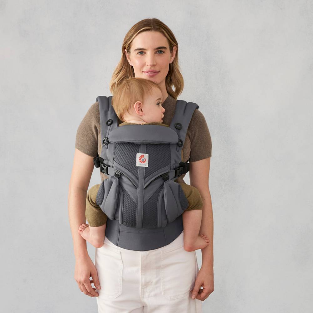 Woman holding a baby in a gray ergonomic carrier against a plain background