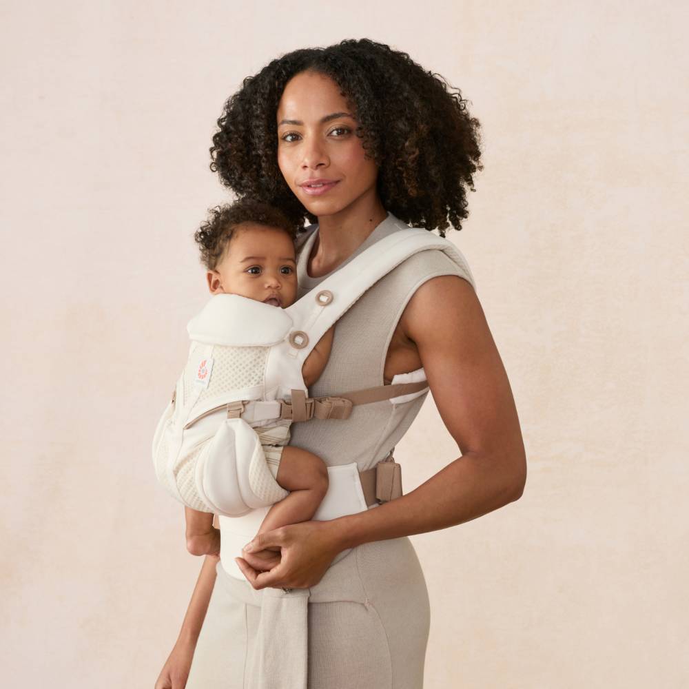 A woman holds a baby in a white and beige front baby carrier against a soft beige studio background.