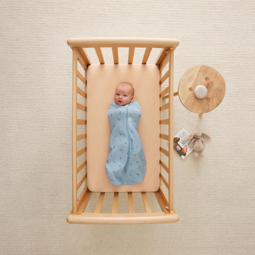 Baby in a blue swaddle blanket in a wooden crib with a beige wall background