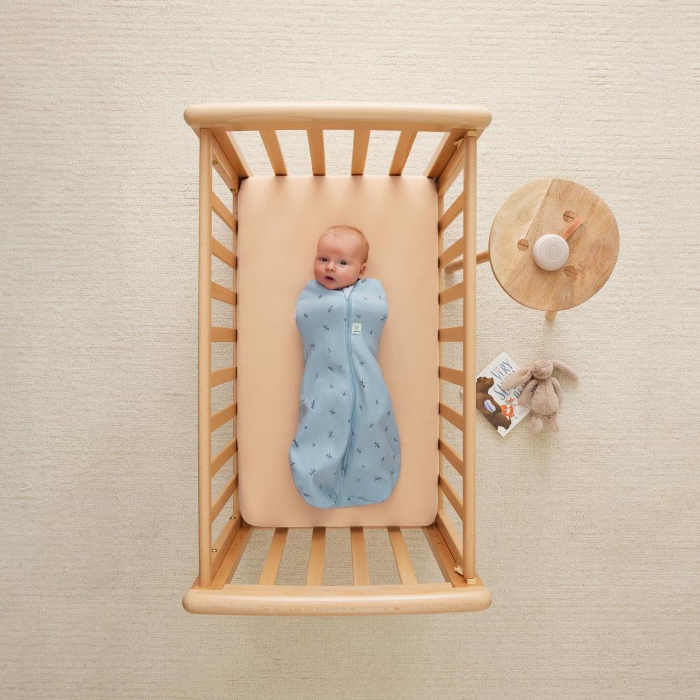 Baby in a blue swaddle blanket in a wooden crib with a beige wall background