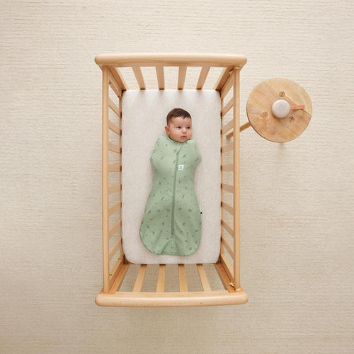 Baby in a green zip swaddle lying in a wooden crib with a beige wall background