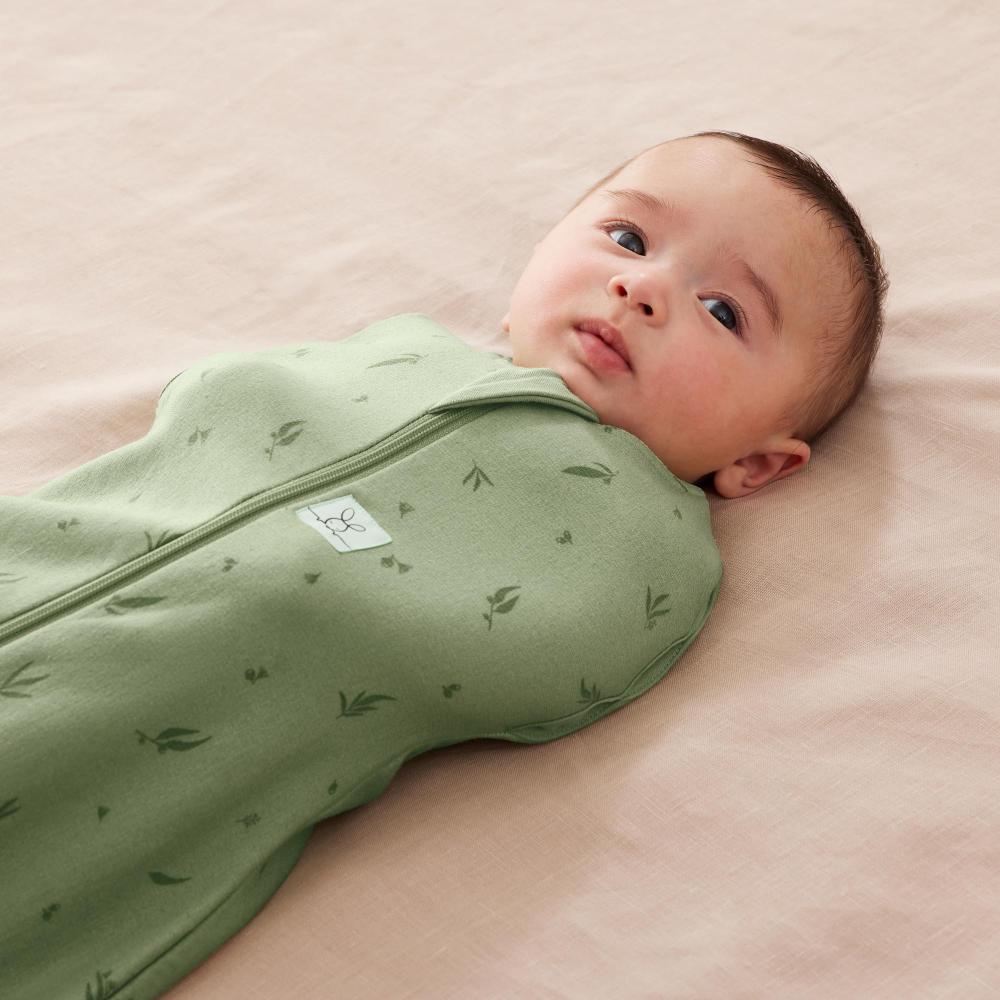 Baby swaddled in a green sleeping cocoon with a pink background