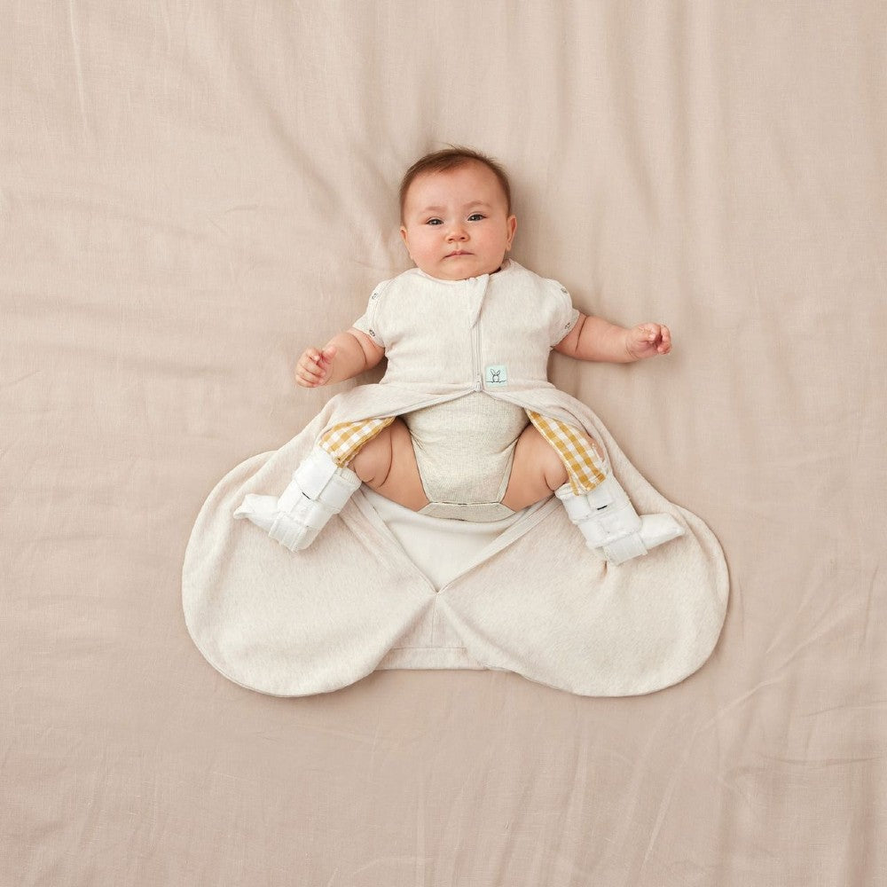 Baby in a beige baby sleeping bag for hip dysplasia with hip[ brace on a beige blanket