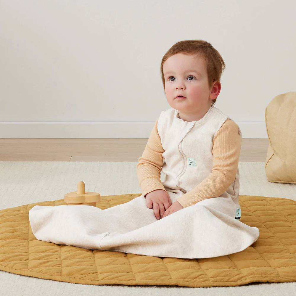 Child sitting on a yellow mat with a white blanket, wearing a beige outfit.