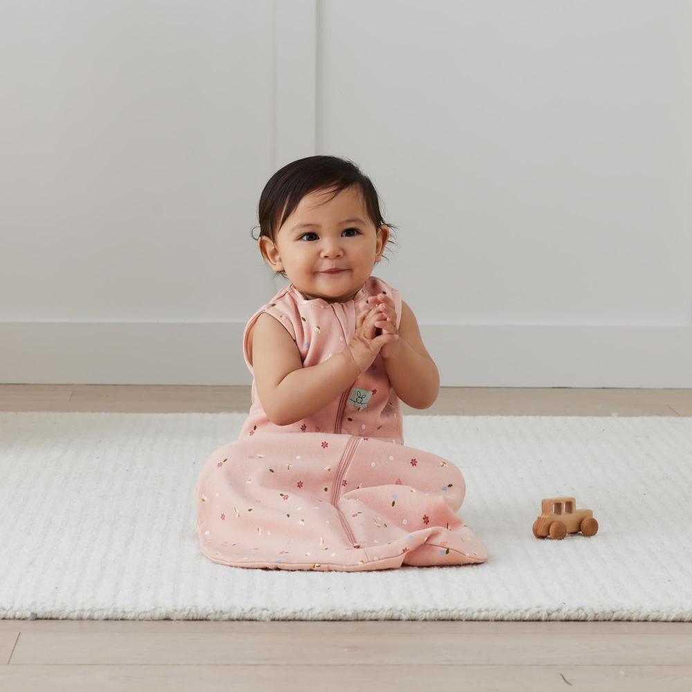 Baby in a pink sleeping bag sitting on a white mat with a wooden toy car.